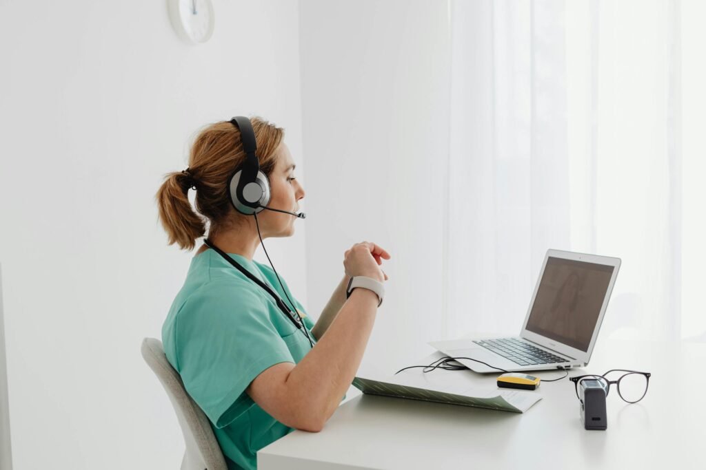 A female doctor using a laptop for an online consultation, wearing a headset in a bright office.